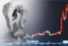 A hand places a small model house in front of a blurry background showing a red and green candlestick chart, symbolizing real estate investment or housing market trends.