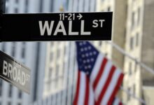 Street signs for Wall Street and Broad Street in New York City with a blurred American flag in the background.