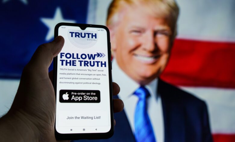 A person holds a phone displaying the Truth Social app with “Follow the Truth” on the screen, in front of a blurred image of Donald Trump and a U.S. flag in the background.