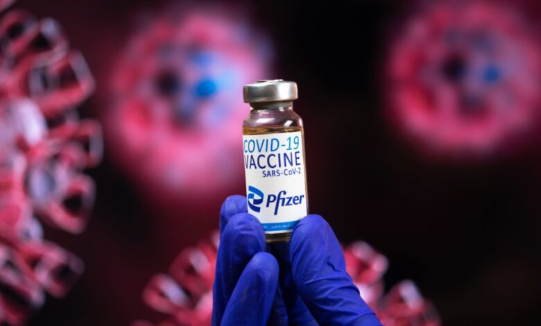 A gloved hand holding a Pfizer COVID-19 vaccine vial with blurred virus particles in the background.
