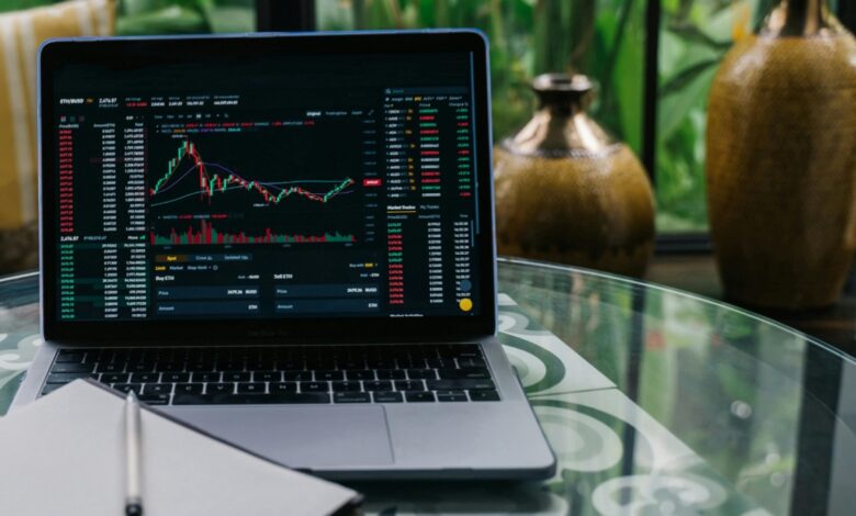 Stock market trading chart on a laptop screen showing market analysis, placed on a glass table with a notebook and pen.