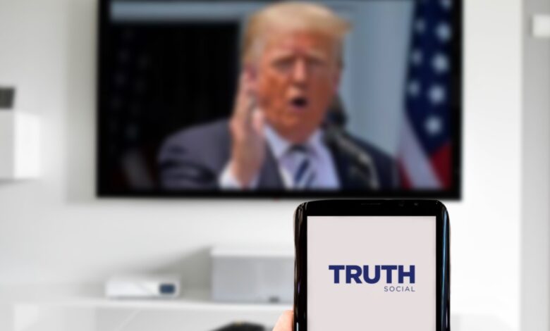 A person holds a smartphone displaying the Truth Social app logo, with a television in the background showing a blurred image of a man speaking in front of American flags.