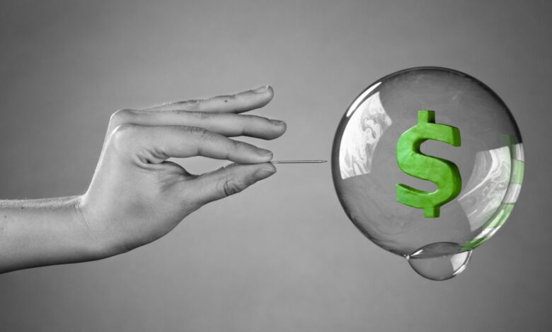 A hand holding a pin approaches a transparent bubble with a green dollar sign inside, symbolizing the potential bursting of a financial bubble.
