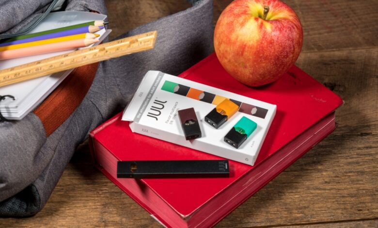 A black Juul vape and pack of nicotine pods sit on top of red school book with an apple on it, next to a grey backpack.