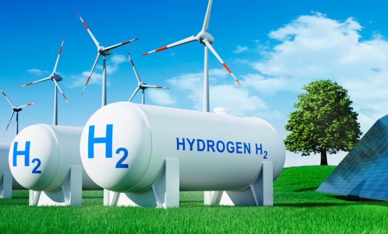 Three white clean hydrogen fuel tanks on green grass next to black solar panels, with windmills in the background.