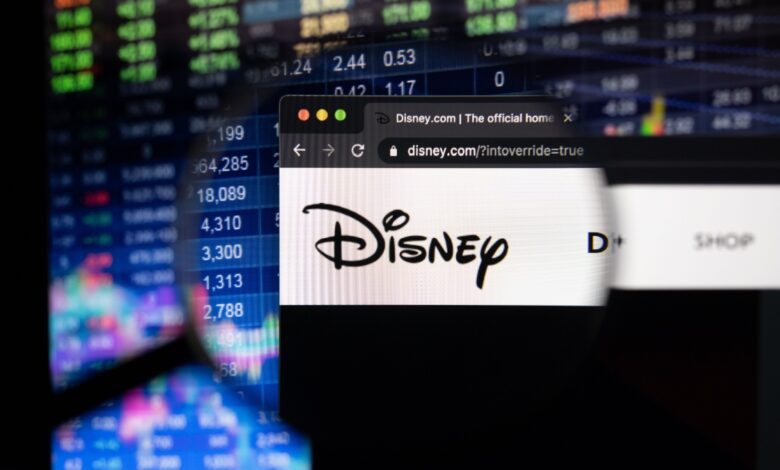 Close-up of Disney.com website viewed through a magnifying glass with Disney logo visible, overlaid on a background of colorful stock market data, representing Disney's online presence, media business performance, and financial market analysis.