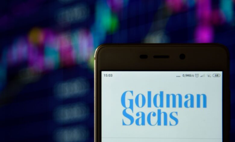 Smartphone screen displaying the Goldman Sachs logo with a blurred background of colorful financial charts, representing investment banking, stock market analytics, global finance, and financial services technology.