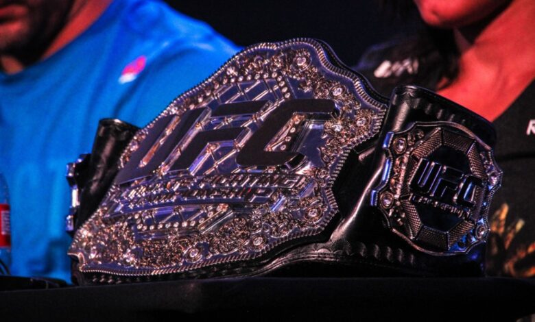 Close-up of the UFC championship belt with intricate silver detailing, displayed during a press event with athletes in the background.