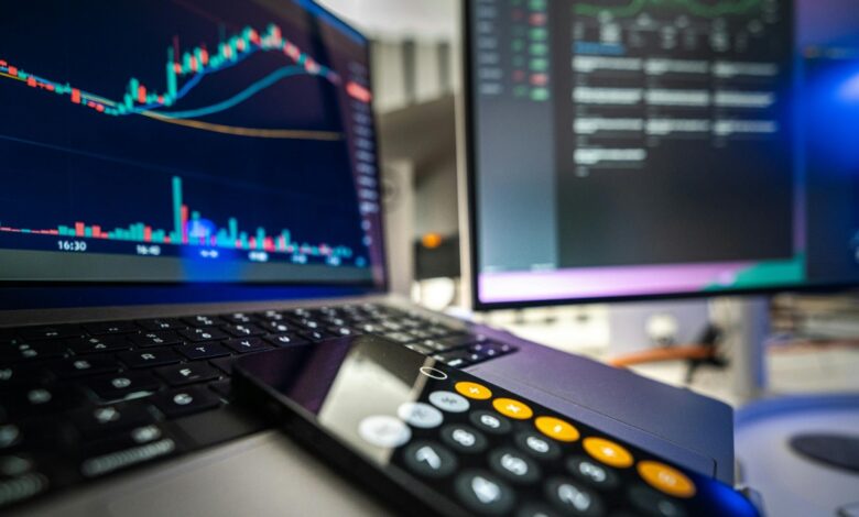 A close-up of a laptop and monitor displaying financial charts and data, with a smartphone showing a calculator app resting on the keyboard. The scene suggests stock trading or financial analysis.
