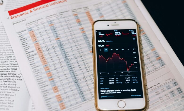 A smartphone displaying an Apple stock chart rests on financial newspapers filled with economic data, tables, and charts. The phone screen shows the stock price trending downward.