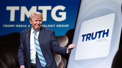 Donald Trump standing in front of a Trump Media and Technology Group sign while pointing to a Truth Social app icon on an enlarged smart phone off to the right.