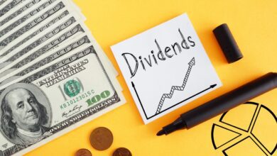 The word dividends written in black marker on a white post-it note, next to several spread out hundred dollar bills on a yellow background.
