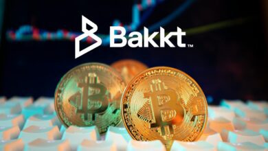 Three Bitcoin on computer keyboard, with white Bakkt logo on dark blue stock chart background.