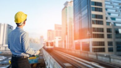 Construction worker wearing a yellow hard hat and holding blueprints, standing on an elevated platform overlooking a train track and modern city skyline with high-rise buildings during sunset.