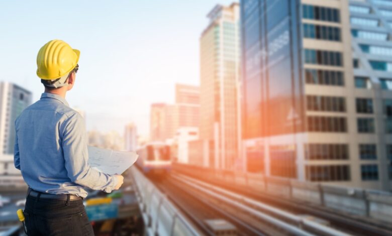 Construction worker wearing a yellow hard hat and holding blueprints, standing on an elevated platform overlooking a train track and modern city skyline with high-rise buildings during sunset.