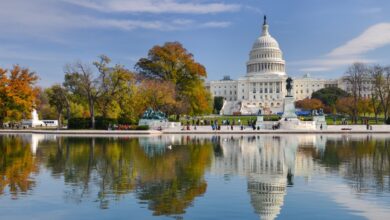 The United States Capitol building in Washington, D.C., viewed from across the reflecting pool on a clear autumn day, with colorful fall trees and visitors walking nearby.