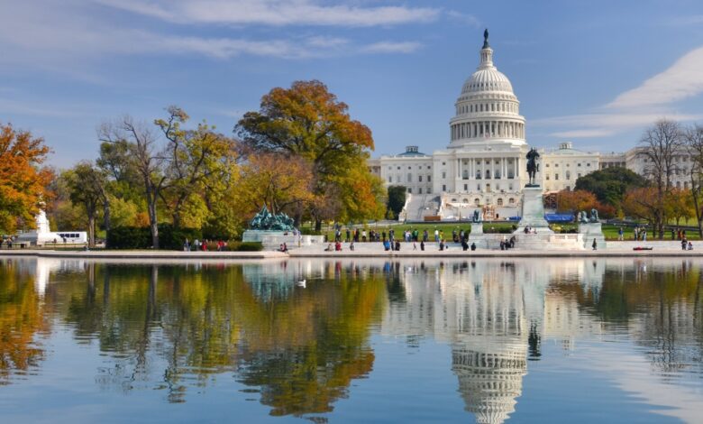 The United States Capitol building in Washington, D.C., viewed from across the reflecting pool on a clear autumn day, with colorful fall trees and visitors walking nearby.