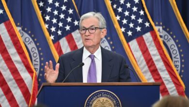 Federal Reserve Chair Jerome Powell in a suit and purple tie speaks at a podium with the seal of the Board of Governors in front. He is gesturing with one hand and standing in front of several American flags and blue banners with gold trim.