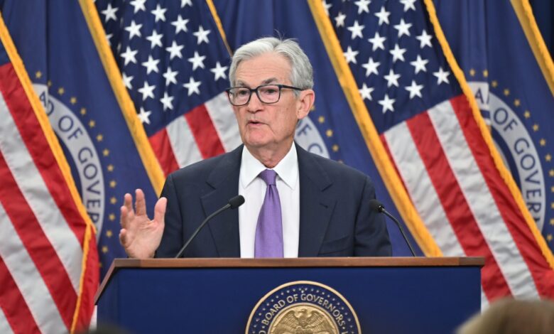Federal Reserve Chair Jerome Powell in a suit and purple tie speaks at a podium with the seal of the Board of Governors in front. He is gesturing with one hand and standing in front of several American flags and blue banners with gold trim.