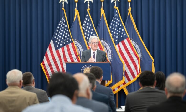 Federal Reserve Chair Jerome Powell speaks at a podium in front of multiple U.S. flags and Federal Reserve banners, addressing an audience.