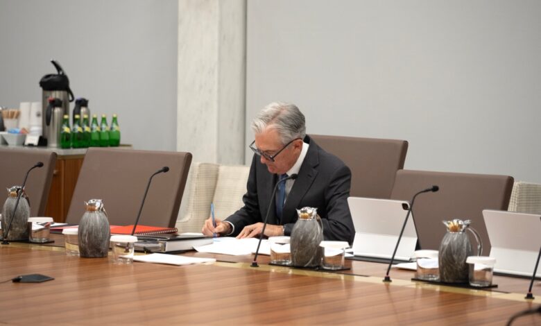 Federal Reserve Chair Jerome Powell sits alone at a conference table, writing on documents. Several microphones, pitchers, and tablets are arranged on the table. In the background, a counter holds bottled water, coffee supplies, and a security camera is mounted on the wall.