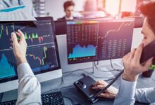 Stock trader on the phone pointing at charts on monitors, with financial graphs and data visible in a busy office.