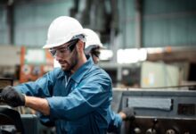 Factory worker in a hard hat and safety glasses operating industrial machinery in a workshop.