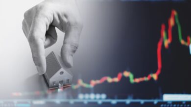 A hand places a small model house in front of a blurry background showing a red and green candlestick chart, symbolizing real estate investment or housing market trends.