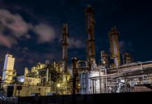 Large industrial oil refinery or chemical plant at night with complex metal structures, tall towers, and bright artificial lighting under a dark sky with clouds.