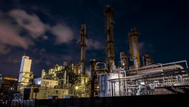 Large industrial oil refinery or chemical plant at night with complex metal structures, tall towers, and bright artificial lighting under a dark sky with clouds.