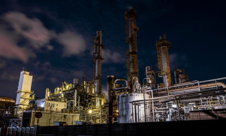 Large industrial oil refinery or chemical plant at night with complex metal structures, tall towers, and bright artificial lighting under a dark sky with clouds.