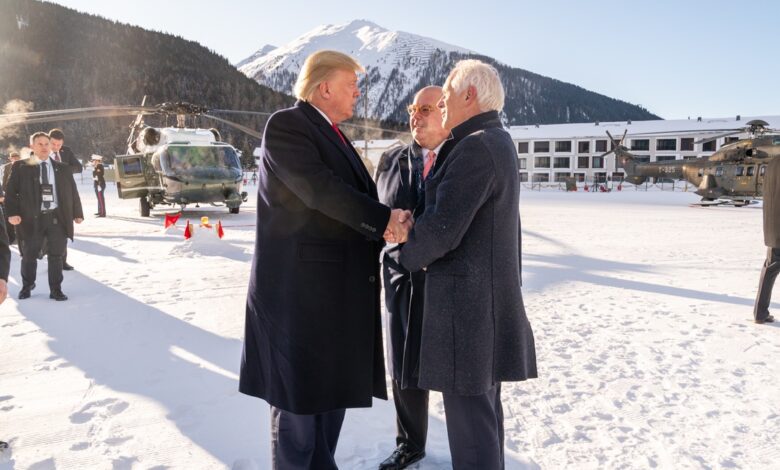 President Donald Trump shaking hands with a man in a snowy mountain setting in Davos, Switzerland, surrounded by officials and helicopters.