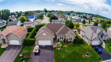 Aerial view of a suburban neighborhood with rows of single-family homes, green lawns, and a playground in the background under a partly cloudy sky.