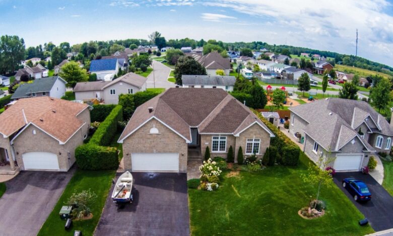 Aerial view of a suburban neighborhood with rows of single-family homes, green lawns, and a playground in the background under a partly cloudy sky.