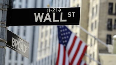 Street signs for Wall Street and Broad Street in New York City with a blurred American flag in the background.