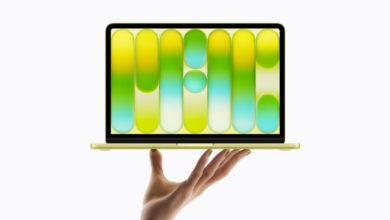 Hand holding a thin yellow laptop against a light gray background, with the open screen displaying abstract rounded vertical shapes in gradient yellow, green, and blue, creating a modern colorful tech product showcase.