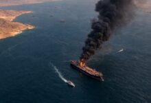 Aerial view of a burning oil tanker with thick black smoke rising above a narrow shipping route in the Straight of Hormuz in the Persian Gulf.